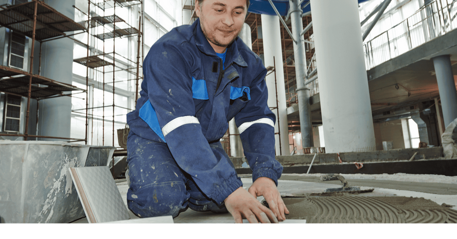 Mitarbeiter in dunkelblauen Arbeitsklamotten verlegt die Fliesen eines Neubaus.